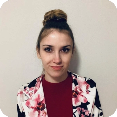 Amelie Meyer, eine junge Frau mit Dutt, trägt einen Blazer mit Blumenmuster und ein rotes Oberteil vor einem neutralen Hintergrund.