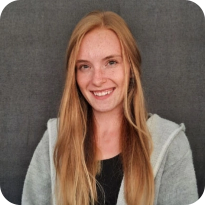 Patricia Jakob, eine junge Frau mit langen blonden Haaren, trägt einen grauen Cardigan und lächelt freundlich vor einem dunklen Hintergrund.