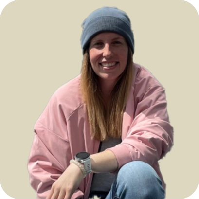 Rebecca Hartmann, eine junge Frau mit blauer Mütze und rosa Jacke, sitzt lächelnd und entspannt vor einem neutralen Hintergrund.