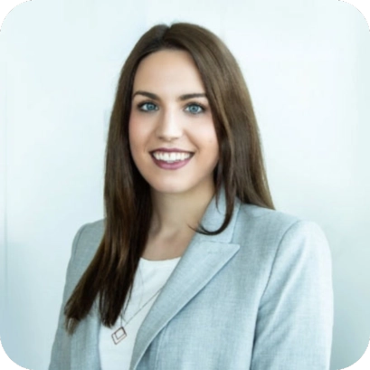 Simone Baumgartner, eine junge Frau in einem grauen Blazer und weißem Oberteil, lächelt professionell vor einem hellen Hintergrund.