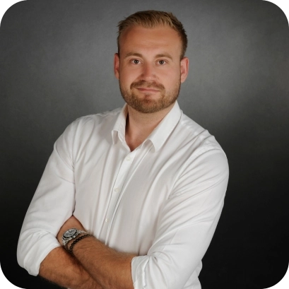 Vincent Karl, ein junger Mann in weißem Hemd mit verschränkten Armen, steht vor einem dunklen Hintergrund und blickt selbstbewusst in die Kamera.