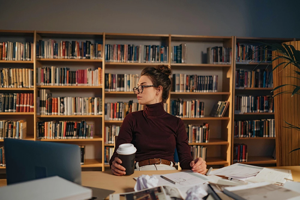 Frau mit Kaffeebecher sitzt in Bibliothek am Schreibtisch – Weiterbildung, Studium oder Meister-BAföG